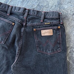 May include: A pair of dark blue denim jeans with red stitching. The jeans have a back pocket with a brown leather patch that says "Wrangler" in white lettering.