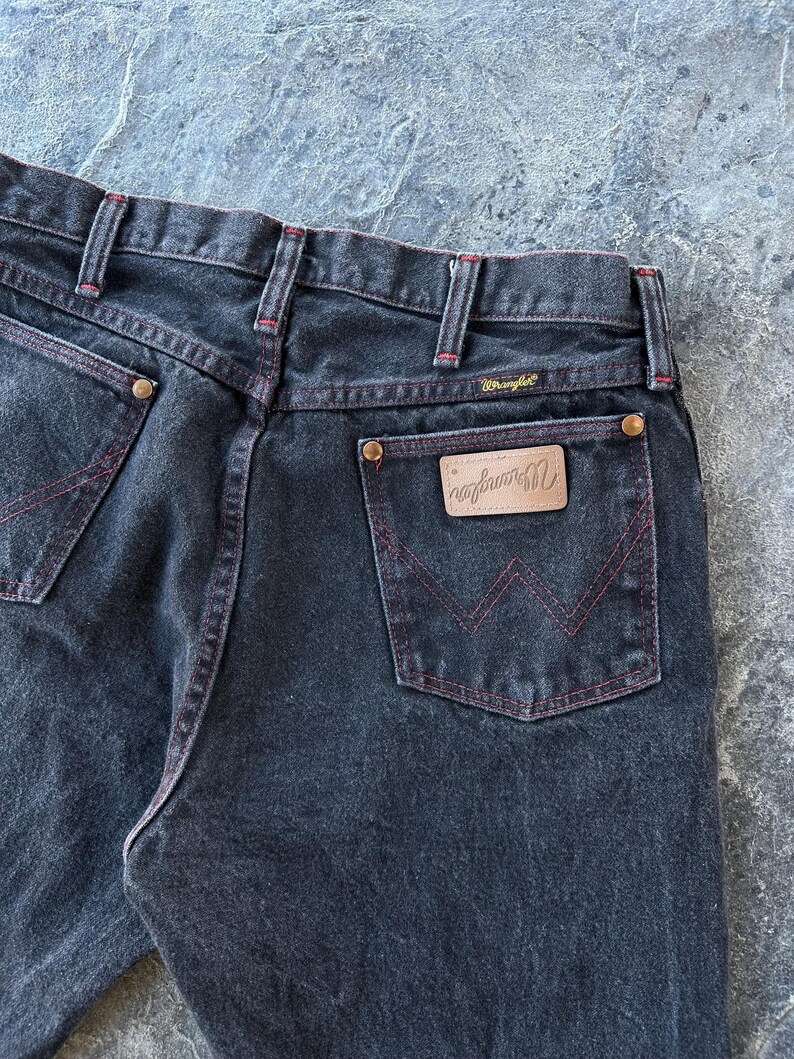 May include: A pair of dark blue denim jeans with red stitching. The jeans have a back pocket with a brown leather patch that says "Wrangler" in white lettering.
