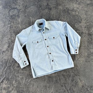 May include: Light blue long-sleeve shirt with two chest pockets and brown buttons. The shirt has a classic collar and buttoned cuffs. The fabric appears to be a soft, woven material.