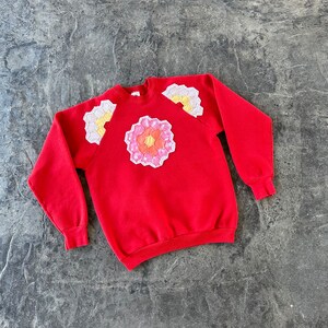 May include: Red sweatshirt with a white and pink flower design on the shoulder.