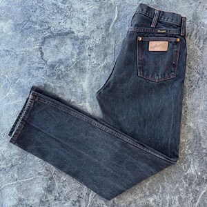 May include: A pair of dark wash denim jeans with red stitching. The jeans have a classic five-pocket style and a Wrangler logo patch on the back pocket.