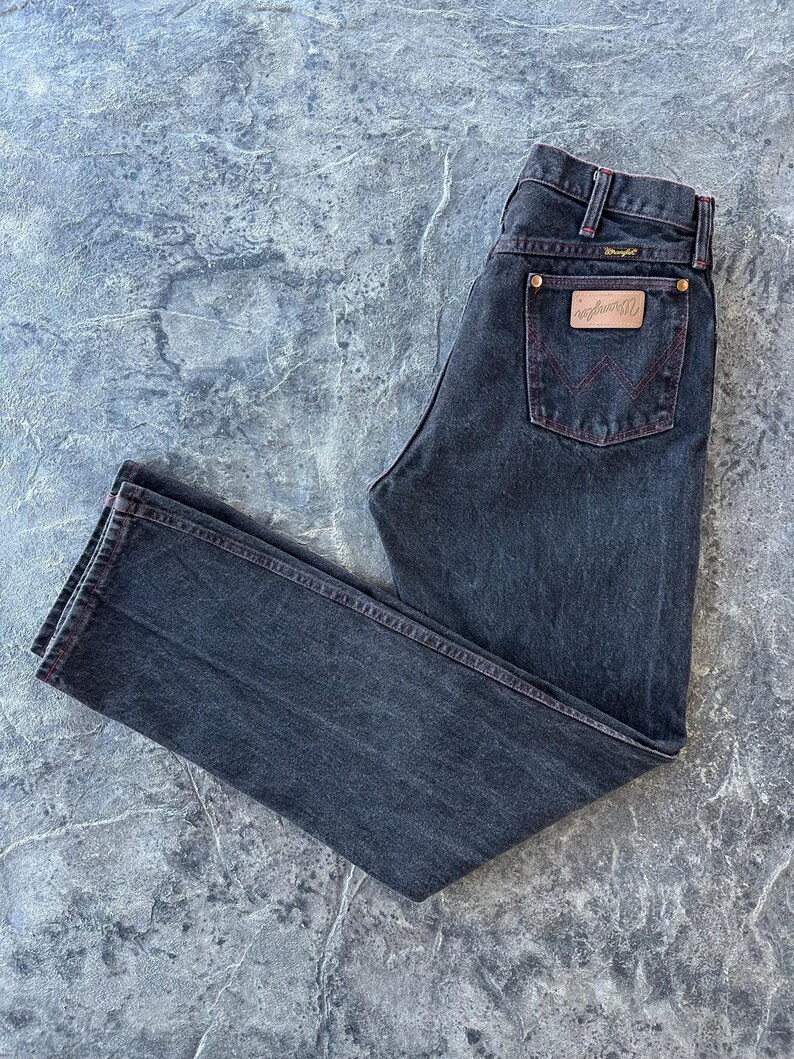 May include: A pair of dark wash denim jeans with red stitching. The jeans have a classic five-pocket style and a Wrangler logo patch on the back pocket.