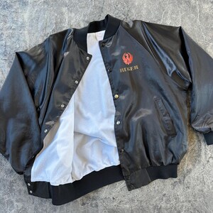 May include: A black satin bomber jacket with a red and gold embroidered logo that says "RUGER". The jacket has a white fleece lining and a snap closure.