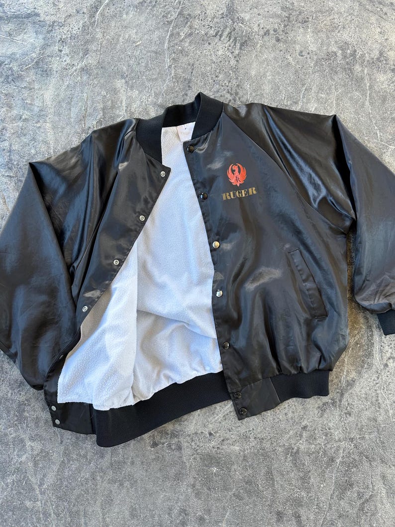 May include: A black satin bomber jacket with a red and gold embroidered logo that says "RUGER". The jacket has a white fleece lining and a snap closure.