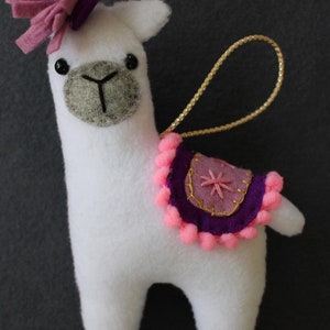 May include: White felt llama ornament with a pink and purple felt flower on its head. The llama has a purple and pink felt saddle with a gold star design and pink pom-poms around the edge. The ornament has a gold string for hanging.