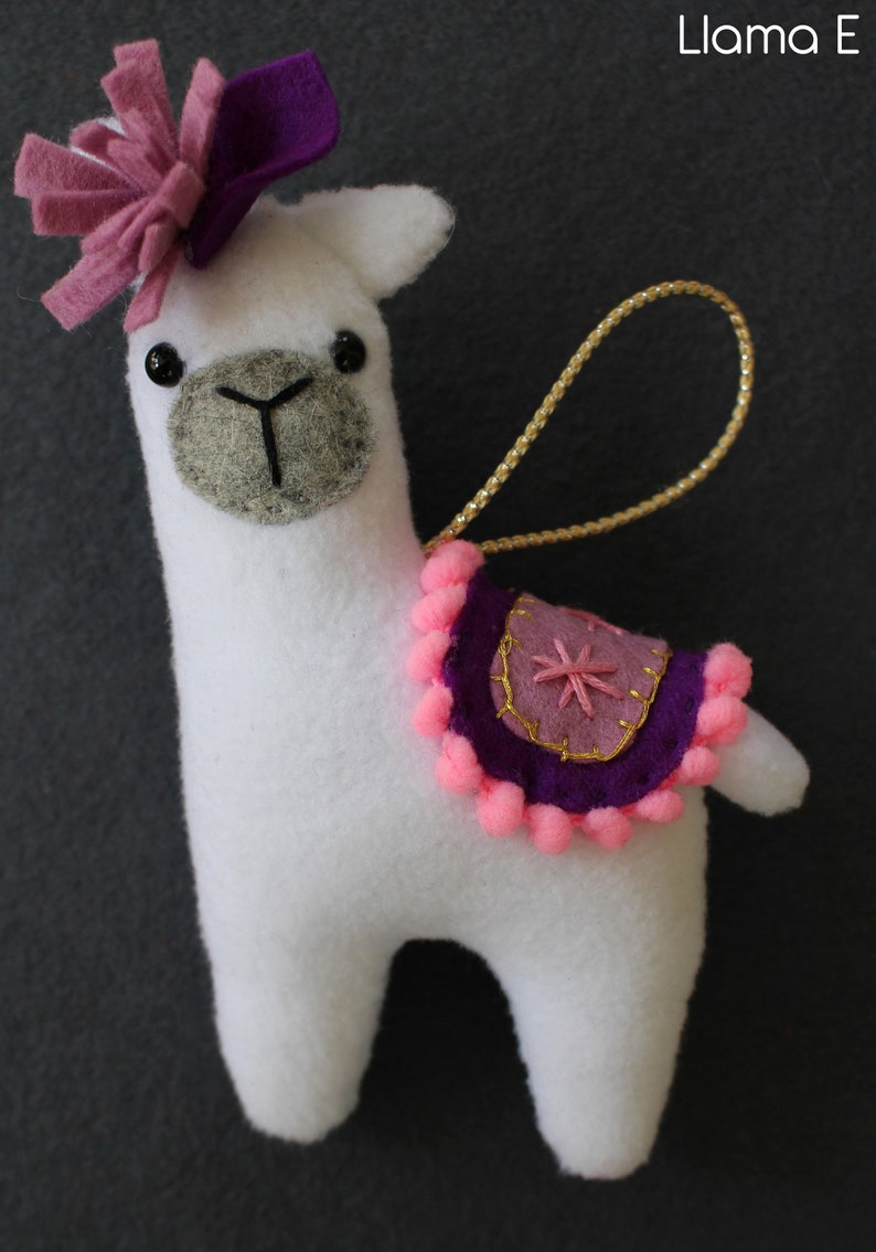 May include: White felt llama ornament with a pink and purple felt flower on its head. The llama has a purple and pink felt saddle with a gold star design and pink pom-poms around the edge. The ornament has a gold string for hanging.