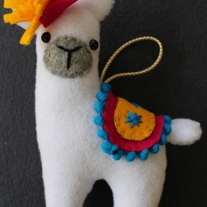 May include: White llama ornament with a yellow flower and red hat. The llama has a blue and pink saddle with yellow accents and blue pom-poms.