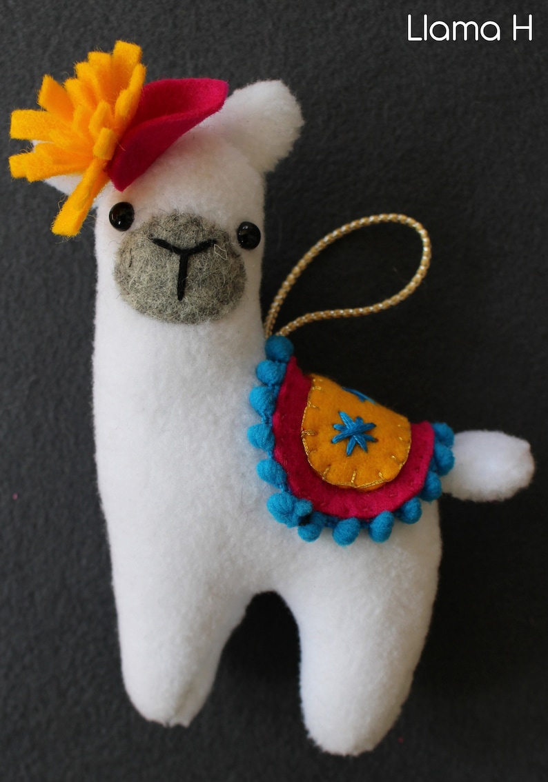 May include: White llama ornament with a yellow flower and red hat. The llama has a blue and pink saddle with yellow accents and blue pom-poms.