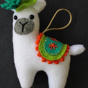 May include: White felt llama ornament with a green felt leaf on its head and a colorful felt saddle with orange pom-poms. The llama has black eyes and a gray felt nose. The ornament has a gold cord for hanging.