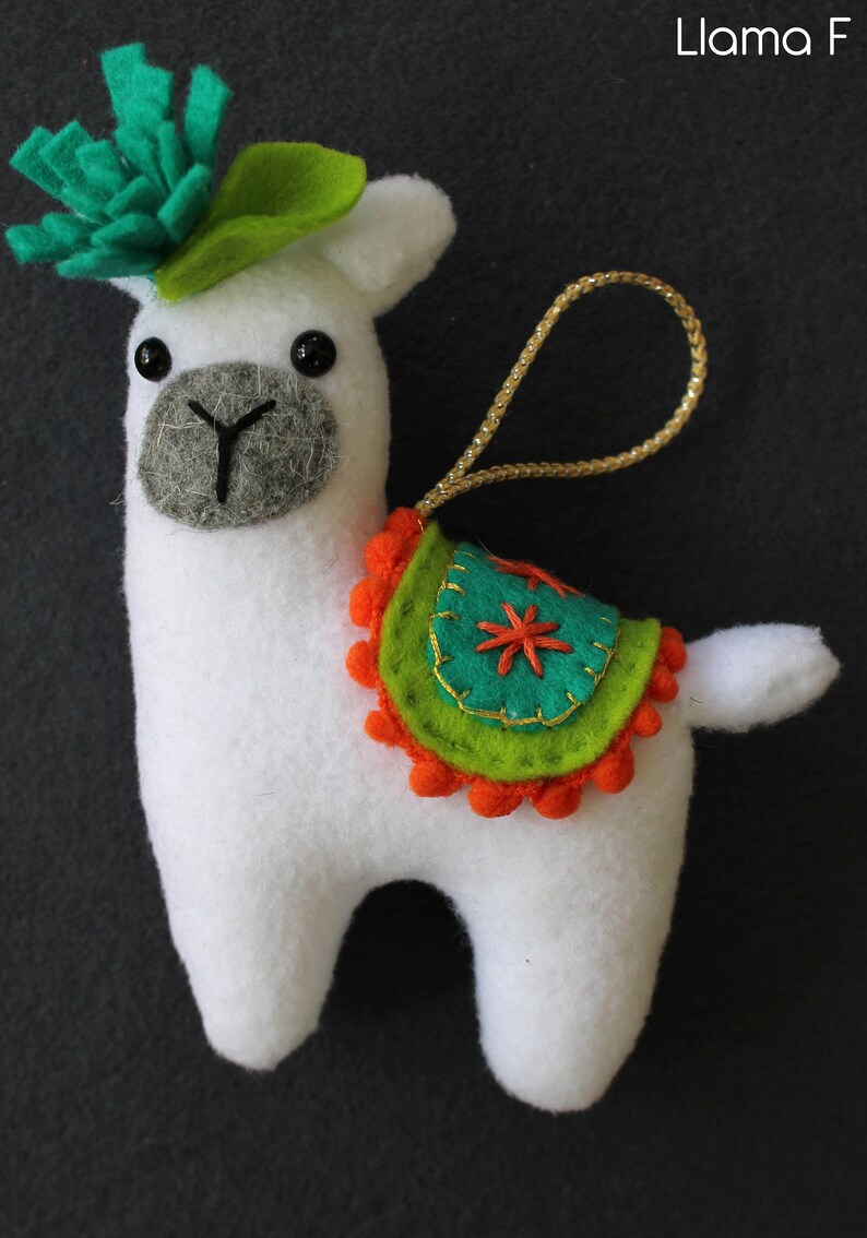 May include: White felt llama ornament with a green felt leaf on its head and a colorful felt saddle with orange pom-poms. The llama has black eyes and a gray felt nose. The ornament has a gold cord for hanging.