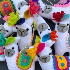 May include: A collection of handmade llama ornaments with colorful felt embellishments. Each llama is white with a different design, including stars, flowers, and geometric patterns.