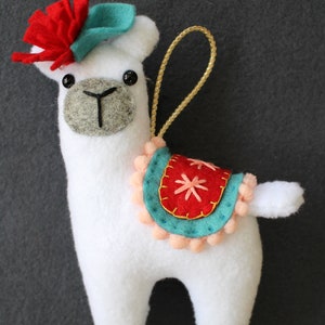 May include: White felt llama ornament with a red felt flower, teal felt hat, and a red and teal felt saddle with a pink pom-pom trim.