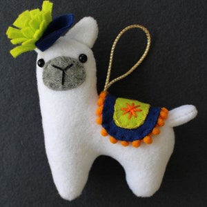 May include: White felt llama ornament with a green felt flower on its head, a blue felt hat, and a blue and green felt saddle with orange pom-poms.