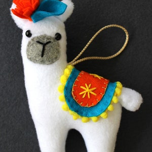May include: White felt llama ornament with a blue felt hat, orange felt flower, and a colorful saddle with yellow pom-poms. The llama has black eyes and a gray felt nose.