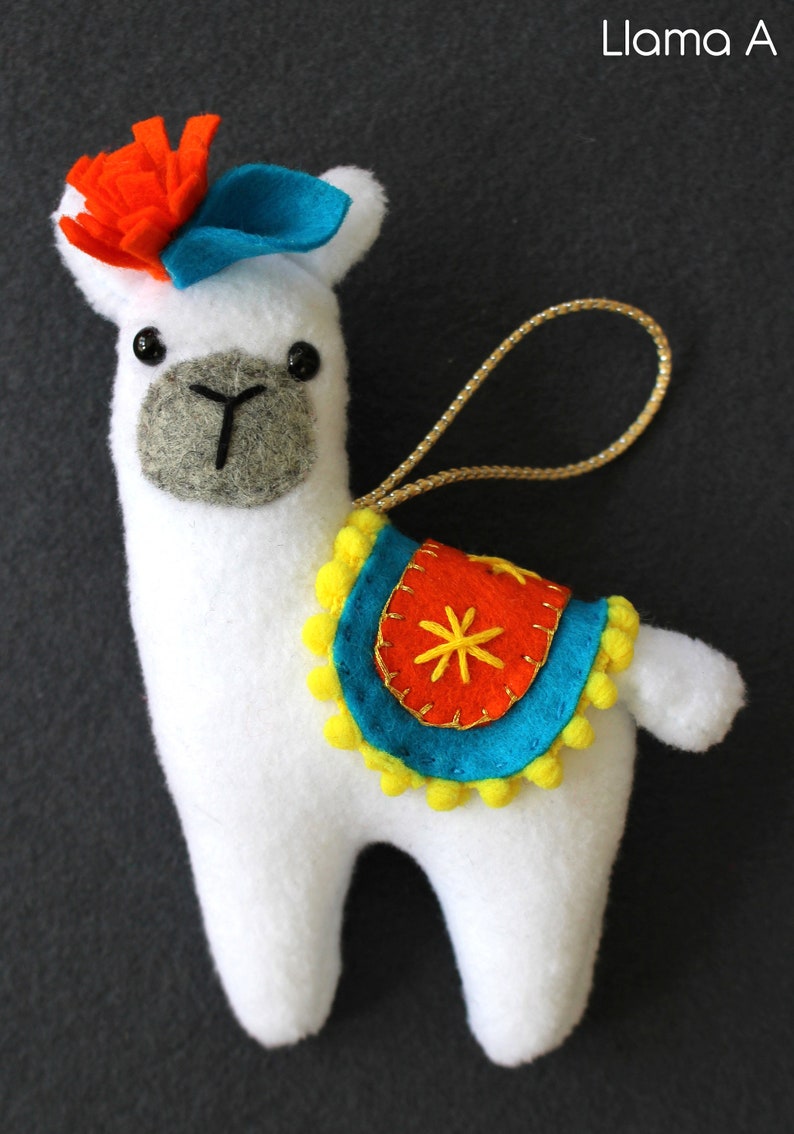 May include: White felt llama ornament with a blue felt hat, orange felt flower, and a colorful saddle with yellow pom-poms. The llama has black eyes and a gray felt nose.