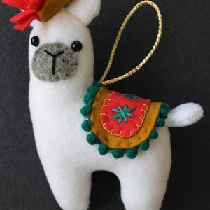 May include: White felt llama ornament with a red and orange felt flower on its head. The llama has a green and gold felt saddle with a red felt flower and green pom-poms around the edge.