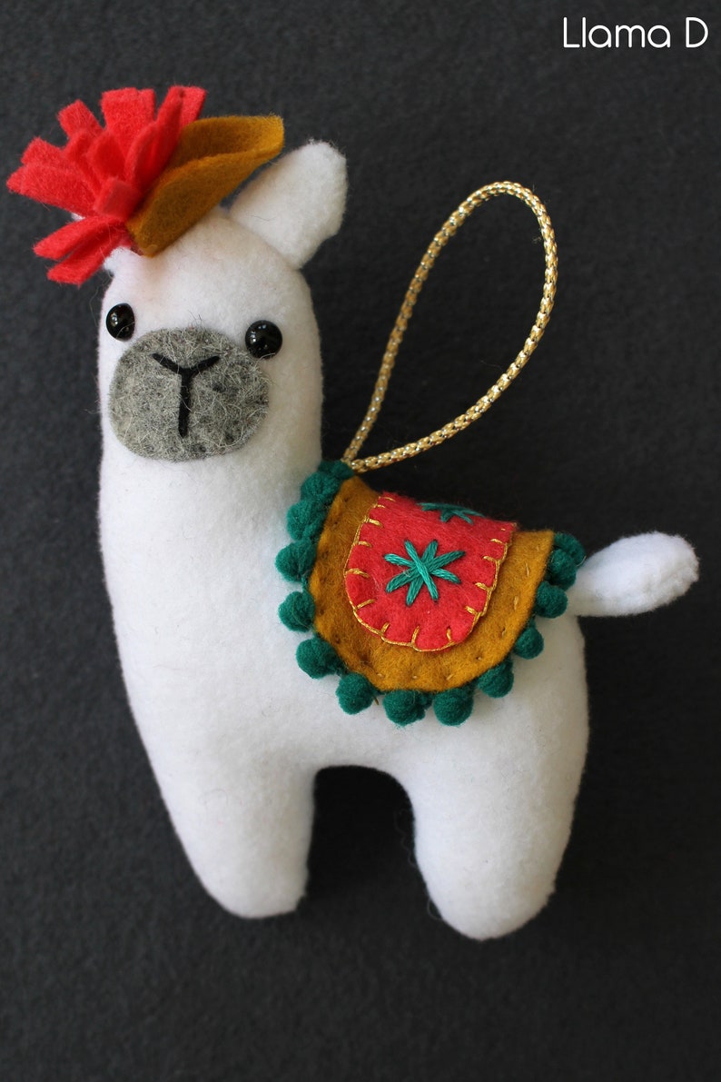 May include: White felt llama ornament with a red and orange felt flower on its head. The llama has a green and gold felt saddle with a red felt flower and green pom-poms around the edge.