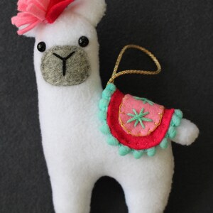 May include: White felt llama ornament with a pink flower on its head and a pink and turquoise saddle with a gold star design. The llama has black eyes and a gray felt nose.
