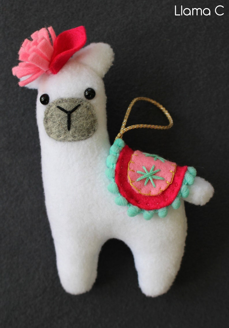 May include: White felt llama ornament with a pink flower on its head and a pink and turquoise saddle with a gold star design. The llama has black eyes and a gray felt nose.