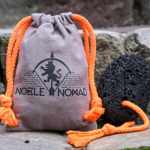 May include: A light brown fabric drawstring pouch with bright orange rope cords. The pouch features a black printed logo of a lion on an arrow with the text NOBLE NOMAD. Beside the pouch is a porous black volcanic rock with an orange rope cord.
