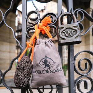Puede incluir: Una bolsa de tela gris con cord&oacute;n que presenta un logotipo negro impreso de un le&oacute;n con una flecha y el texto NOBLE NOMAD, unida a una cuerda naranja brillante con una piedra p&oacute;mez porosa de color marr&oacute;n oscuro, colgada de una verja de metal.