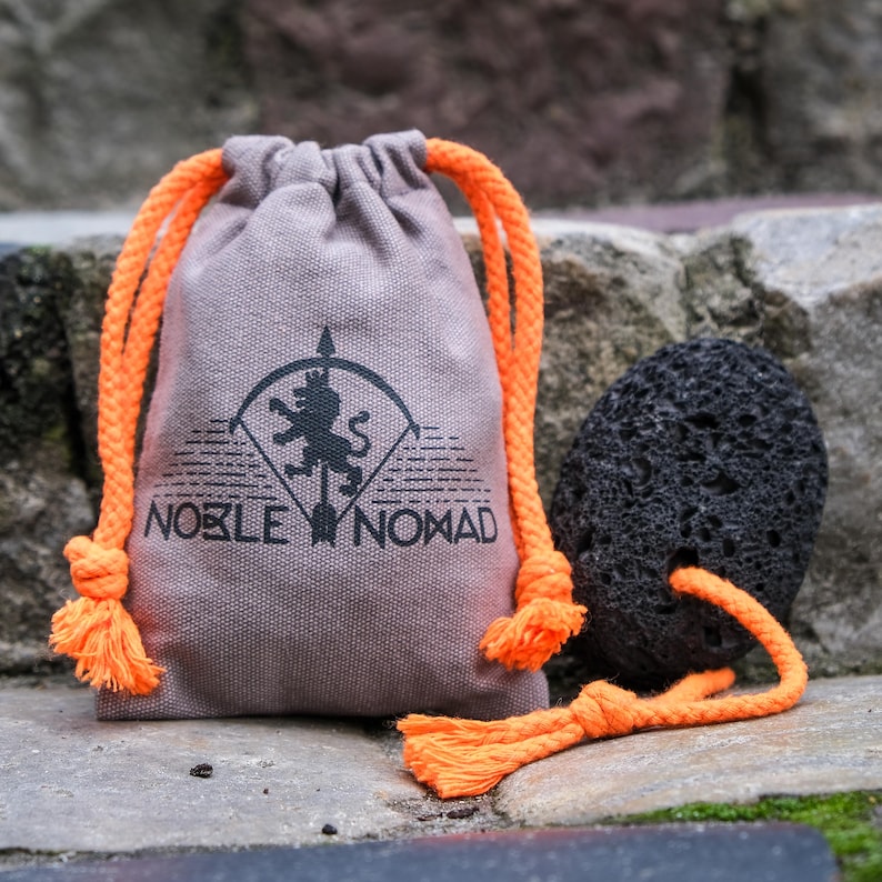 May include: A light brown fabric drawstring pouch with bright orange rope cords. The pouch features a black printed logo of a lion on an arrow with the text NOBLE NOMAD. Beside the pouch is a porous black volcanic rock with an orange rope cord.