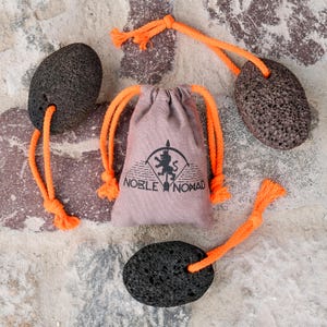 Puede incluir: Tres piedras p&oacute;mez ovaladas, de color gris oscuro y porosas, cada una unida a una cuerda de color naranja brillante, dispuestas alrededor de una peque&ntilde;a bolsa de tela de color marr&oacute;n claro con cord&oacute;n y un logo negro con Noble Nomad.