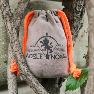 Puede incluir: Una bolsa de tela de color marr&oacute;n claro con cierre de cord&oacute;n que presenta un estampado gr&aacute;fico negro de un le&oacute;n sobre una flecha dentro de un arco, con el texto NOBLE NOMAD debajo. La bolsa tiene cordones trenzados de color naranja.