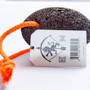 Puede incluir: Una piedra p&oacute;mez ovalada, porosa y de color gris oscuro con un lazo de cuerda trenzada de color naranja brillante. Incluye una etiqueta rectangular blanca con un gr&aacute;fico negro de un le&oacute;n con una flecha, s&iacute;mbolos, c&oacute;digo y NOBLENOMAD.CO.