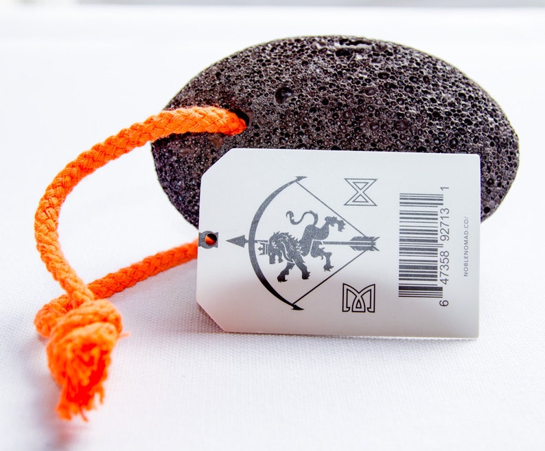 Puede incluir: Una piedra p&oacute;mez ovalada, porosa y de color gris oscuro con un lazo de cuerda trenzada de color naranja brillante. Incluye una etiqueta rectangular blanca con un gr&aacute;fico negro de un le&oacute;n con una flecha, s&iacute;mbolos, c&oacute;digo y NOBLENOMAD.CO.