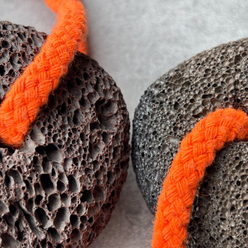 Puede incluir: Dos piedras p&oacute;mez porosas de color gris oscuro, cada una con un cord&oacute;n trenzado de color naranja brillante enhebrado por el centro. Las piedras tienen una textura rugosa e irregular con numerosos orificios peque&ntilde;os en la superficie, lo que contrasta con el cord&oacute;n liso.