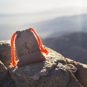 Puede incluir: Una bolsa con cord&oacute;n de color topo con cuerdas y borlas de color naranja brillante. La parte frontal presenta un logotipo negro impreso de una silueta de le&oacute;n dentro de un arco circular, con el texto NOBLE NOMAD impreso bajo el dise&ntilde;o.