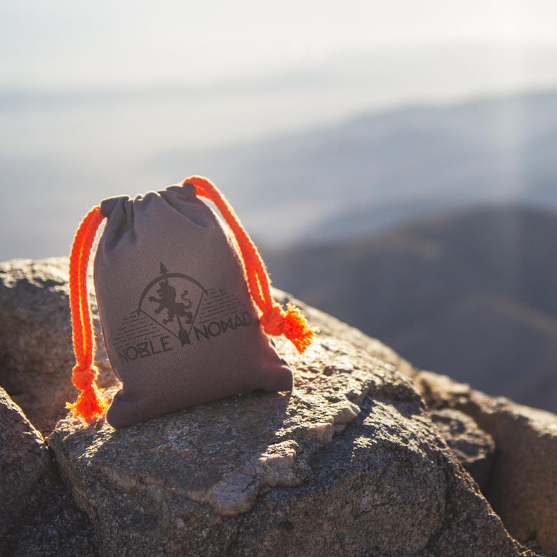 Puede incluir: Una bolsa con cord&oacute;n de color topo con cuerdas y borlas de color naranja brillante. La parte frontal presenta un logotipo negro impreso de una silueta de le&oacute;n dentro de un arco circular, con el texto NOBLE NOMAD impreso bajo el dise&ntilde;o.