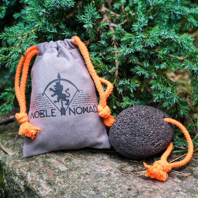 May include: A light brown canvas drawstring pouch with bright orange cords and a black printed logo featuring a lion and the text NOBLE NOMAD, accompanied by a dark gray porous volcanic pumice stone with an attached orange cord loop.