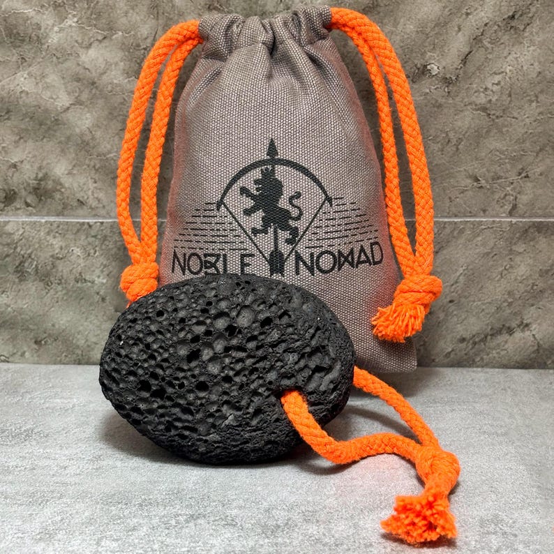 Puede incluir: Una piedra p&oacute;mez negra, porosa y de forma ovalada con una cuerda de color naranja brillante enhebrada, situada frente a una bolsa beige con cord&oacute;n. La bolsa presenta un logotipo negro impreso de un le&oacute;n sobre una flecha con el texto NOBLE NOMAD.