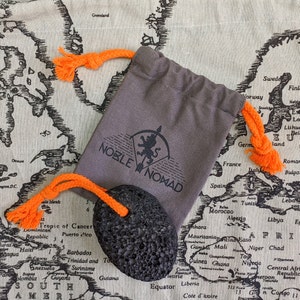 Puede incluir: Una bolsa de tela gris con cord&oacute;n, con el texto impreso en negro NOBLE NOMAD y un logotipo de le&oacute;n, junto a una piedra volc&aacute;nica negra porosa unida a un cord&oacute;n de cuerda naranja brillante que combina con los lazos de la bolsa.