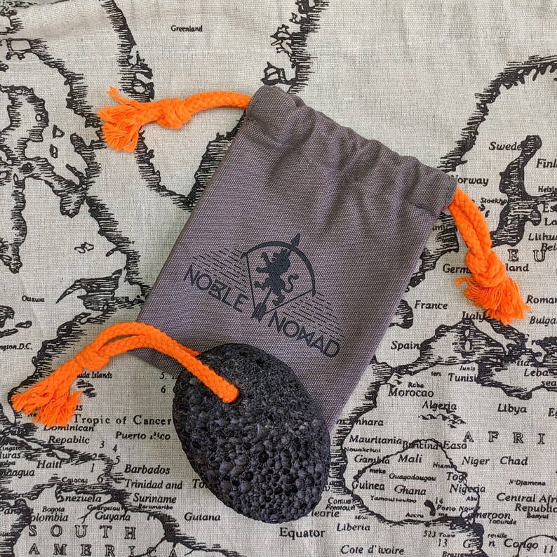 Puede incluir: Una bolsa de tela gris con cord&oacute;n, con el texto impreso en negro NOBLE NOMAD y un logotipo de le&oacute;n, junto a una piedra volc&aacute;nica negra porosa unida a un cord&oacute;n de cuerda naranja brillante que combina con los lazos de la bolsa.