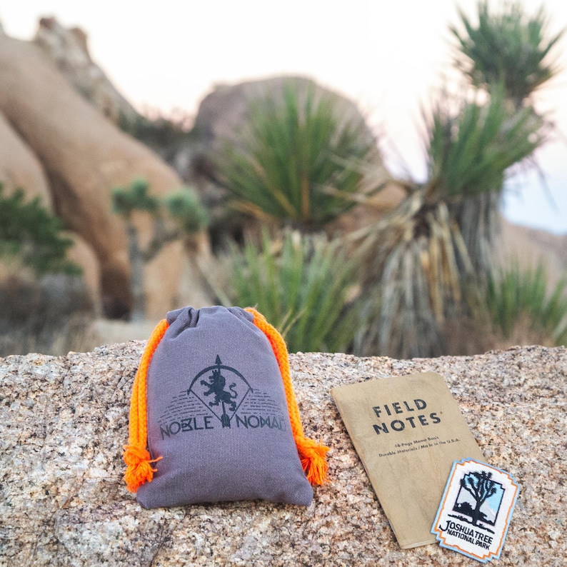 Puede incluir: Una bolsa gris con cord&oacute;n de color naranja brillante que presenta un logotipo negro de un le&oacute;n y el texto Noble Nomad, una libreta Field Notes de color tostado y un parche rectangular del Parque Nacional Joshua Tree con un paisaje.