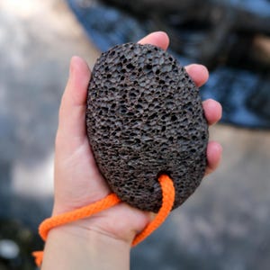 Puede incluir: Una piedra p&oacute;mez de color marr&oacute;n oscuro con forma ovalada y una textura porosa y rugosa. La piedra presenta un peque&ntilde;o orificio cerca del borde inferior, por el que pasa un cord&oacute;n trenzado de color naranja brillante para colgarla.