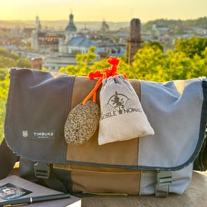 Puede incluir: Un bolso mensajero multicolor con un parche del logotipo de Timbuk2, con un panel central marr&oacute;n y paneles laterales grises. Una piedra gris porosa y una bolsa beige con cord&oacute;n y el texto Noble Nomad est&aacute;n atadas al bolso con un cord&oacute;n naranja.