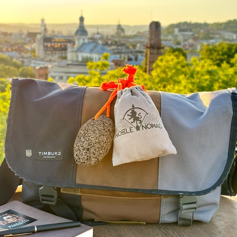 Puede incluir: Un bolso mensajero multicolor con un parche del logotipo de Timbuk2, con un panel central marr&oacute;n y paneles laterales grises. Una piedra gris porosa y una bolsa beige con cord&oacute;n y el texto Noble Nomad est&aacute;n atadas al bolso con un cord&oacute;n naranja.