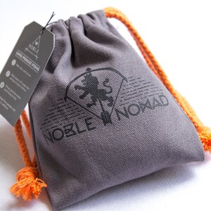 Puede incluir: Una bolsa de lona gris con cord&oacute;n y cuerdas de color naranja brillante. La bolsa presenta un logotipo negro impreso de un le&oacute;n y el texto NOBLE NOMAD. Se adjunta una etiqueta rectangular gris con el texto NOBLE NOMAD LAVA PUMICE STONE.