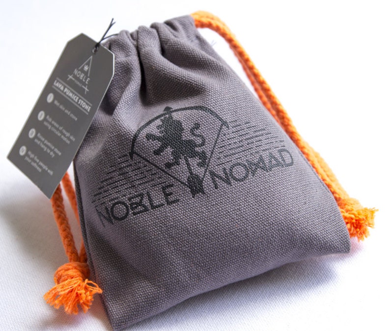 Puede incluir: Una bolsa de lona gris con cord&oacute;n y cuerdas de color naranja brillante. La bolsa presenta un logotipo negro impreso de un le&oacute;n y el texto NOBLE NOMAD. Se adjunta una etiqueta rectangular gris con el texto NOBLE NOMAD LAVA PUMICE STONE.