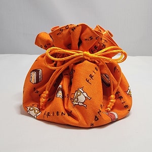Puede incluir: Bolsa de cordón naranja con temática de la serie de televisión Friends. La bolsa está cubierta con un patrón repetido del logotipo de la serie, un pavo y una rebanada de pastel.