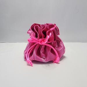 Puede incluir: Una pequeña bolsa rosa con cordón y estampado de cachemira blanco. La bolsa está fruncida en la parte superior con un cordón rosa a juego. La bolsa está sobre una superficie blanca.