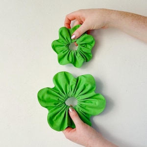 May include: Two green fabric scrunchies in the shape of flowers. The larger scrunchie is being held by a hand.