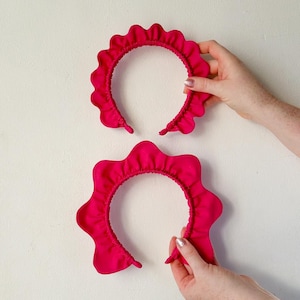 May include: Two vibrant pink headbands with a ruffled edge. The headbands are made of a soft fabric and are held against a white background. The headbands have a wavy, scalloped design.