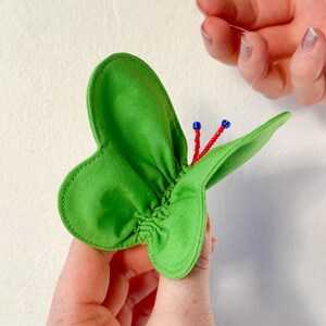 May include: A green fabric butterfly with red and blue beads sewn to the center. The butterfly is being held by a hand.