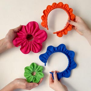 以下が含まれることがあります： 異なる色とスタイルのファブリックヘアスクランチが4つ。スクランチは、フリルのある縁のある花の形をしています。色はピンク、オレンジ、ブルー、グリーンです。