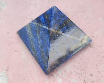 May include: A lapis lazuli pyramid with a square base. The pyramid is a deep blue colour with streaks of white and gold. The pyramid is on a pink surface.